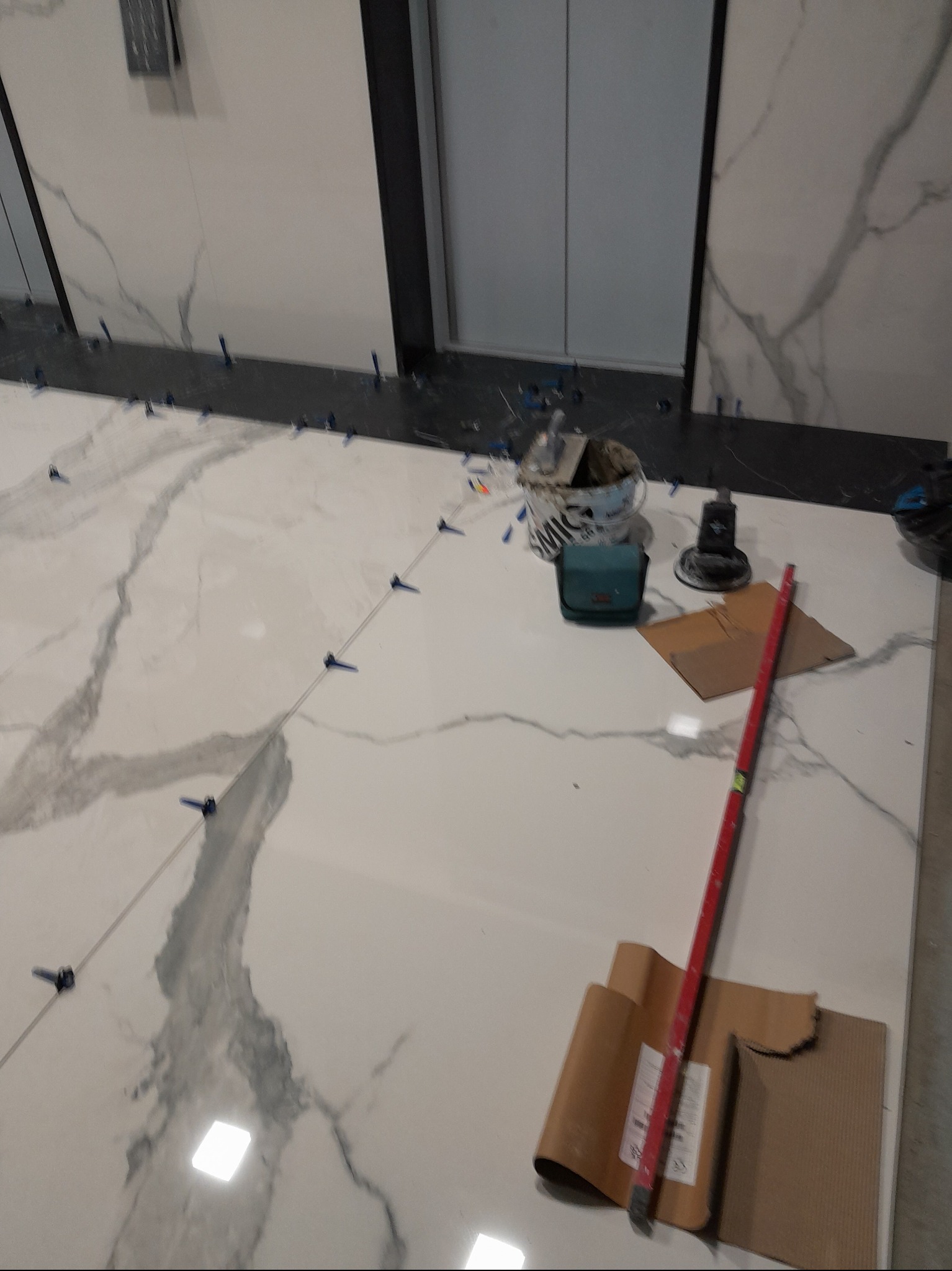 Układanie wielkoformatowych płytek imitujących marmur na podłodze, widoczne narzędzia glazurnicze, system poziomowania płytek i fragment windy.