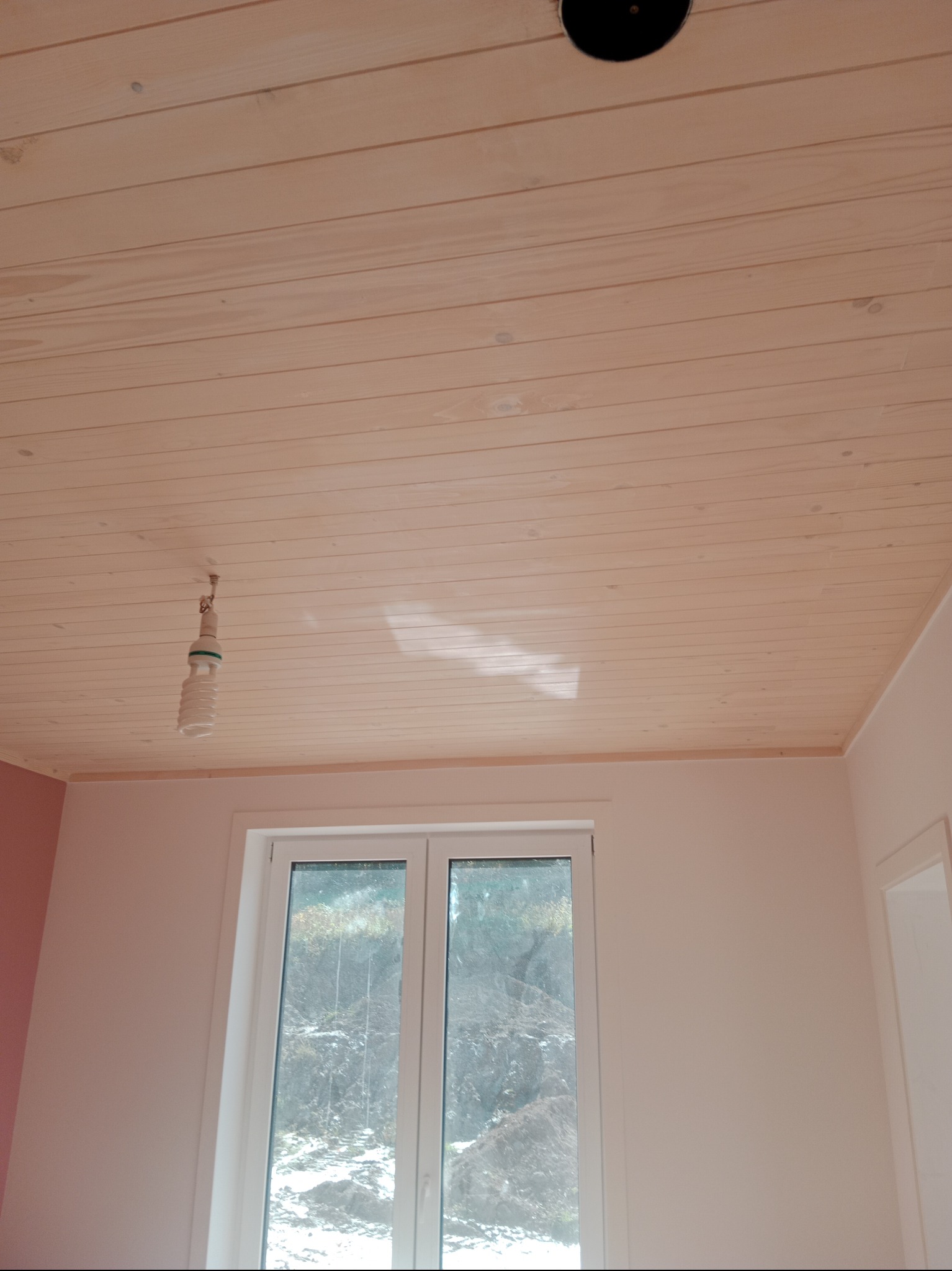 Drewniany sufit z widocznym otworem na oświetlenie i wiszącą żarówką energooszczędną, świeżo wykończone wnętrze z oknem i fragmentem różowej ściany.