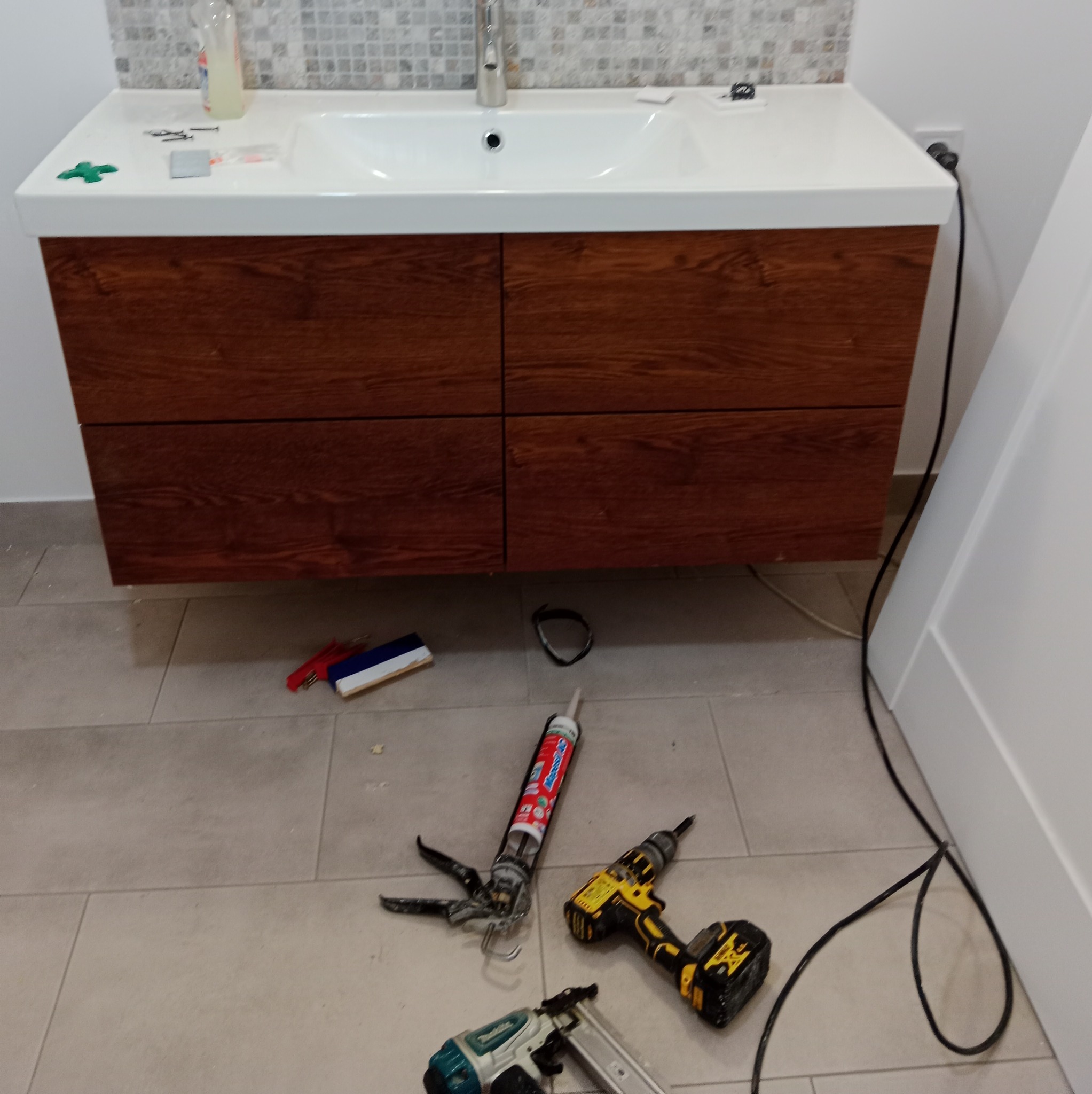 Nowa szafka pod umywalkę z drewnopodobnymi frontami, biały blat i umywalka. Na podłodze narzędzia: pistolet do silikonu, wiertarka, zszywacz pneumatyczny, silikon, poziomica, kabel zasilający.