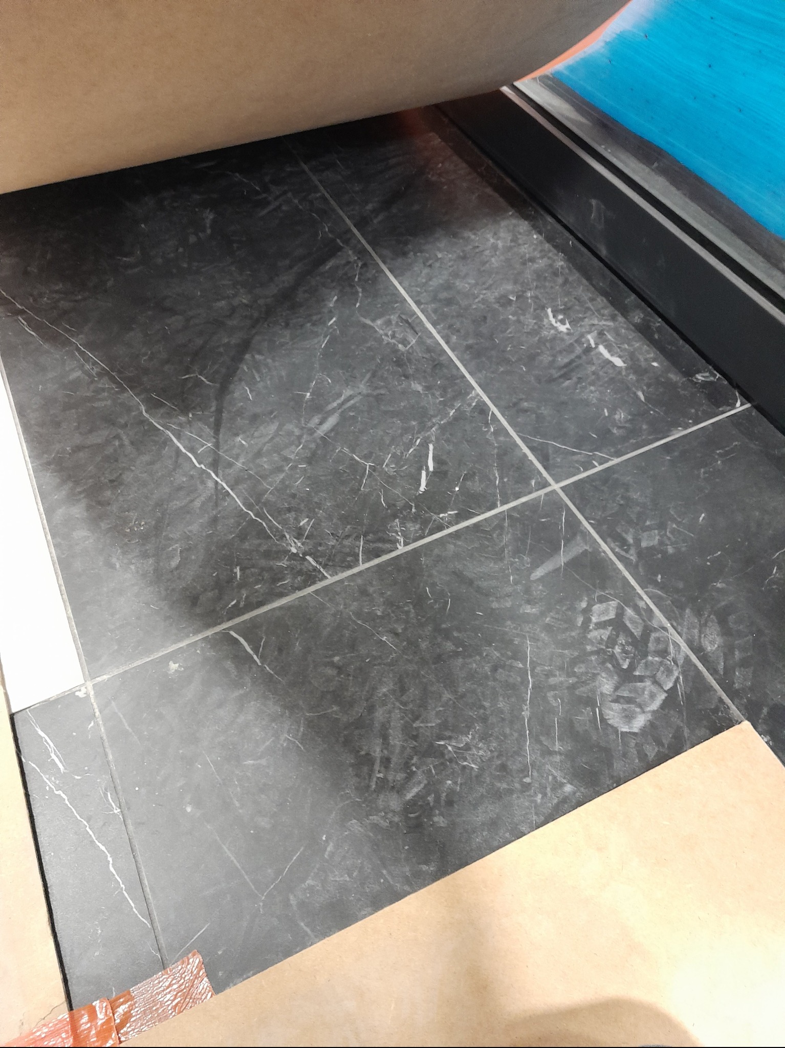 Układanie płytek imitujących czarny marmur z białymi żyłkami, widoczne ślady butów na powierzchni, zabezpieczone kartonem.