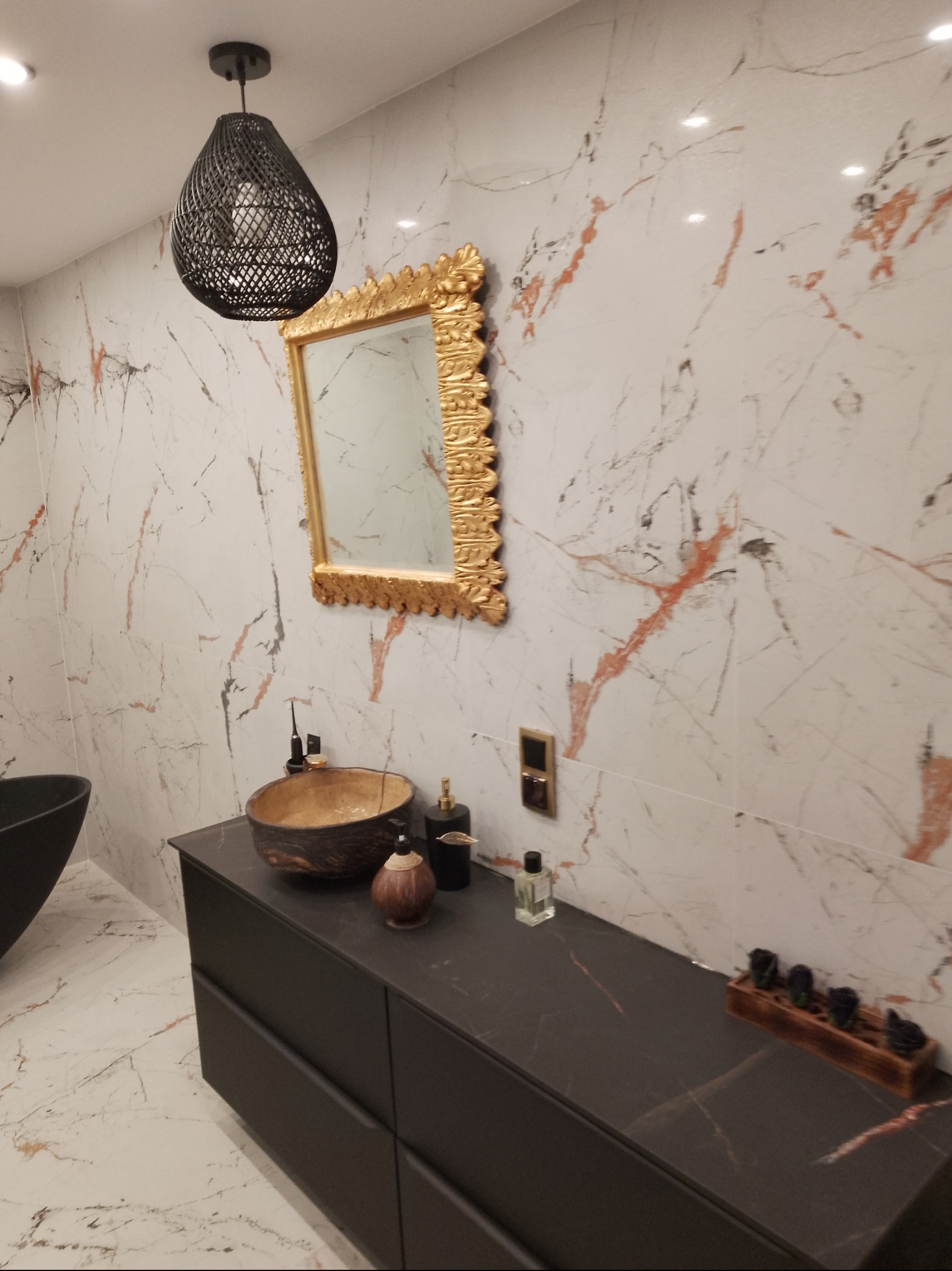 Elegancka łazienka z marmurowymi płytkami, złotym lustrem i designerską umywalką. Czarna szafka pod umywalką i pleciona lampa dodają nowoczesnego charakteru.
