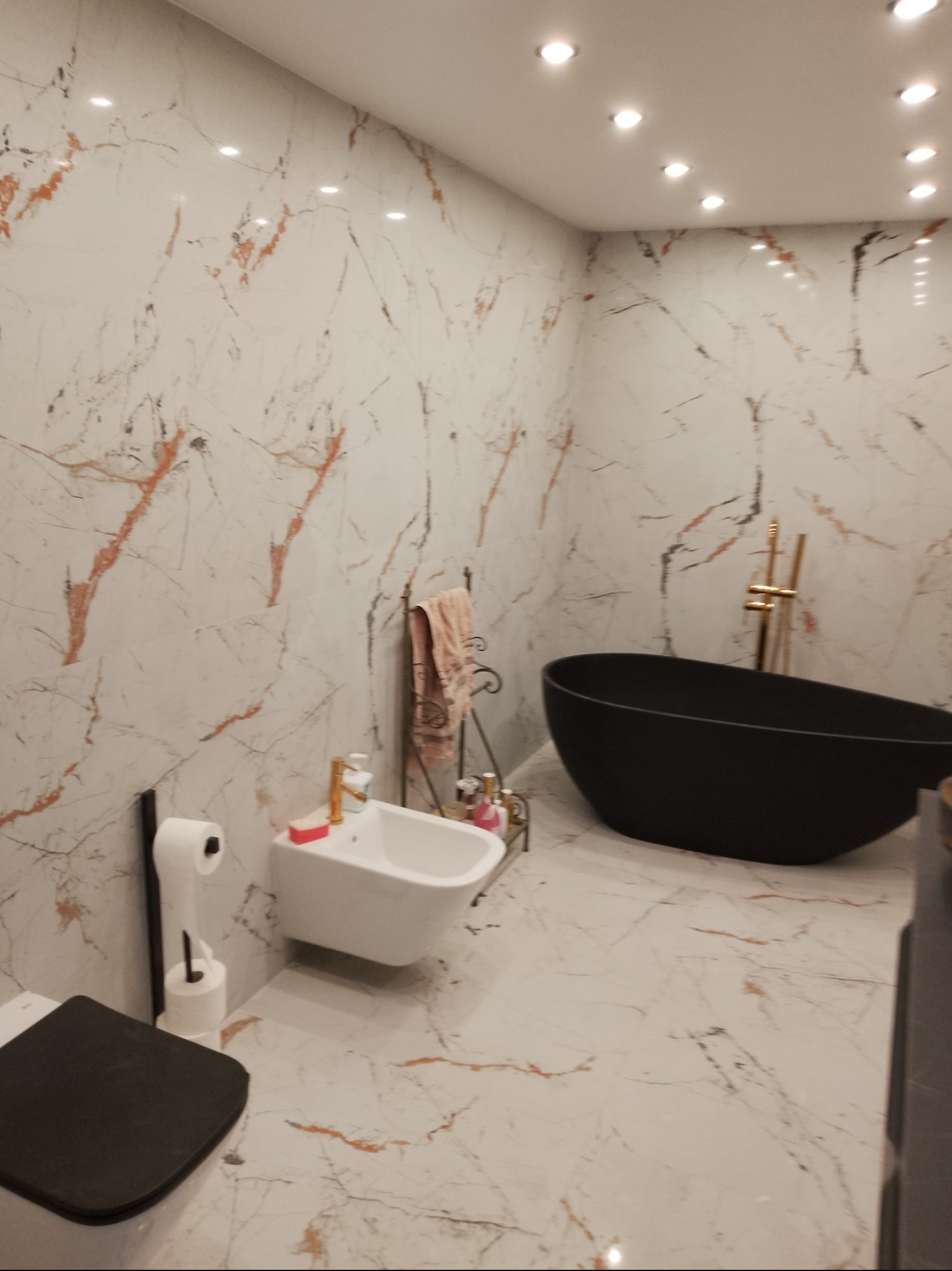 Nowoczesna łazienka z czarną wanną wolnostojącą i marmurowymi płytkami. Bidet i toaleta w jasnych kolorach. Oświetlenie sufitowe punktowe.