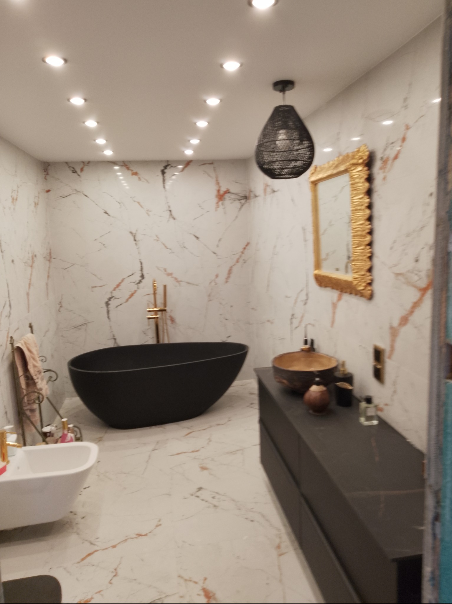 Nowoczesna łazienka z czarną wanną wolnostojącą i umywalką w stylu boho. Marmurowe ściany z delikatnym żyłowaniem, złote akcenty armatury i dekoracyjne oświetlenie.
