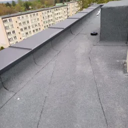 Płaski dach pokryty papą z widocznymi pęknięciami, obróbki blacharskie w kolorze grafitowym, w tle budynki mieszkalne.