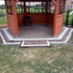 Drewniana altana ogrodowa z brązowym dachem i ścianami, posadowiona na betonowej wylewce otoczonej kostką brukową w odcieniach szarości i beżu, z zielonym trawnikiem w tle.