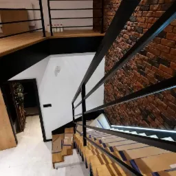 Nowoczesne schody drewniane z czarną metalową balustradą, zabezpieczone kartonem na czas montażu, widok z góry na ceglaną ścianę w tle.