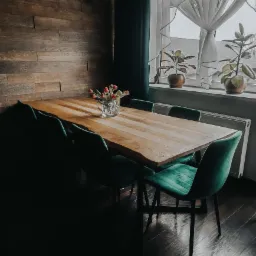 Drewniany stół z naturalną krawędzią blatu, otoczony zielonymi, welurowymi krzesłami, na tle drewnianej ściany i okna z roślinami doniczkowymi.