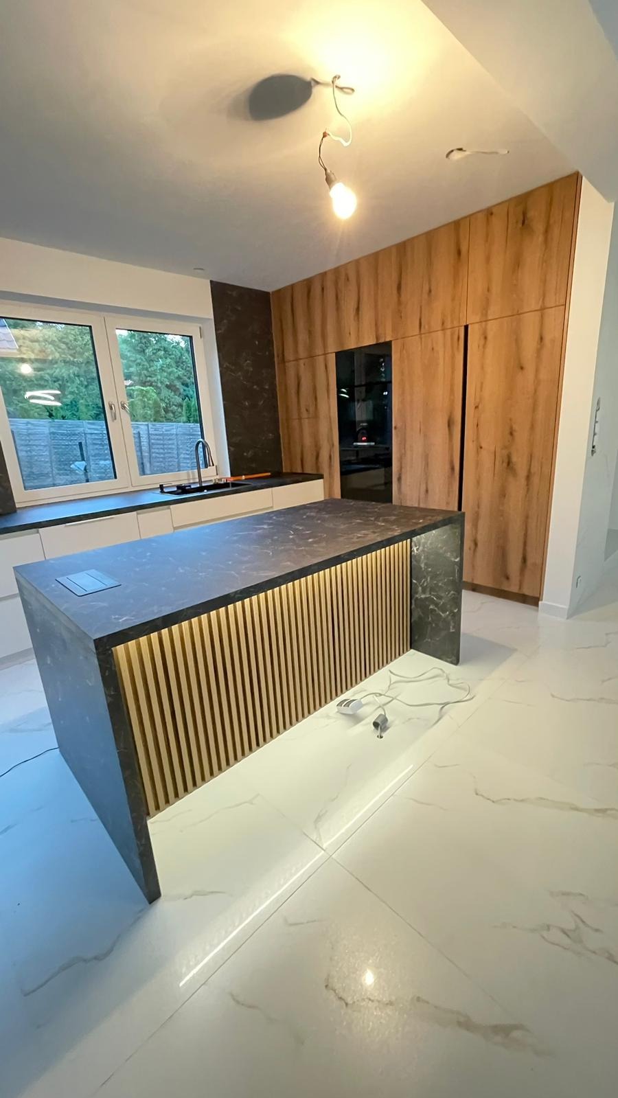 Nowoczesna wyspa kuchenna z blatem imitującym marmur, podświetlonymi drewnianymi lamelami i białą podstawą, na tle drewnianych szafek i okna z widokiem na ogród.