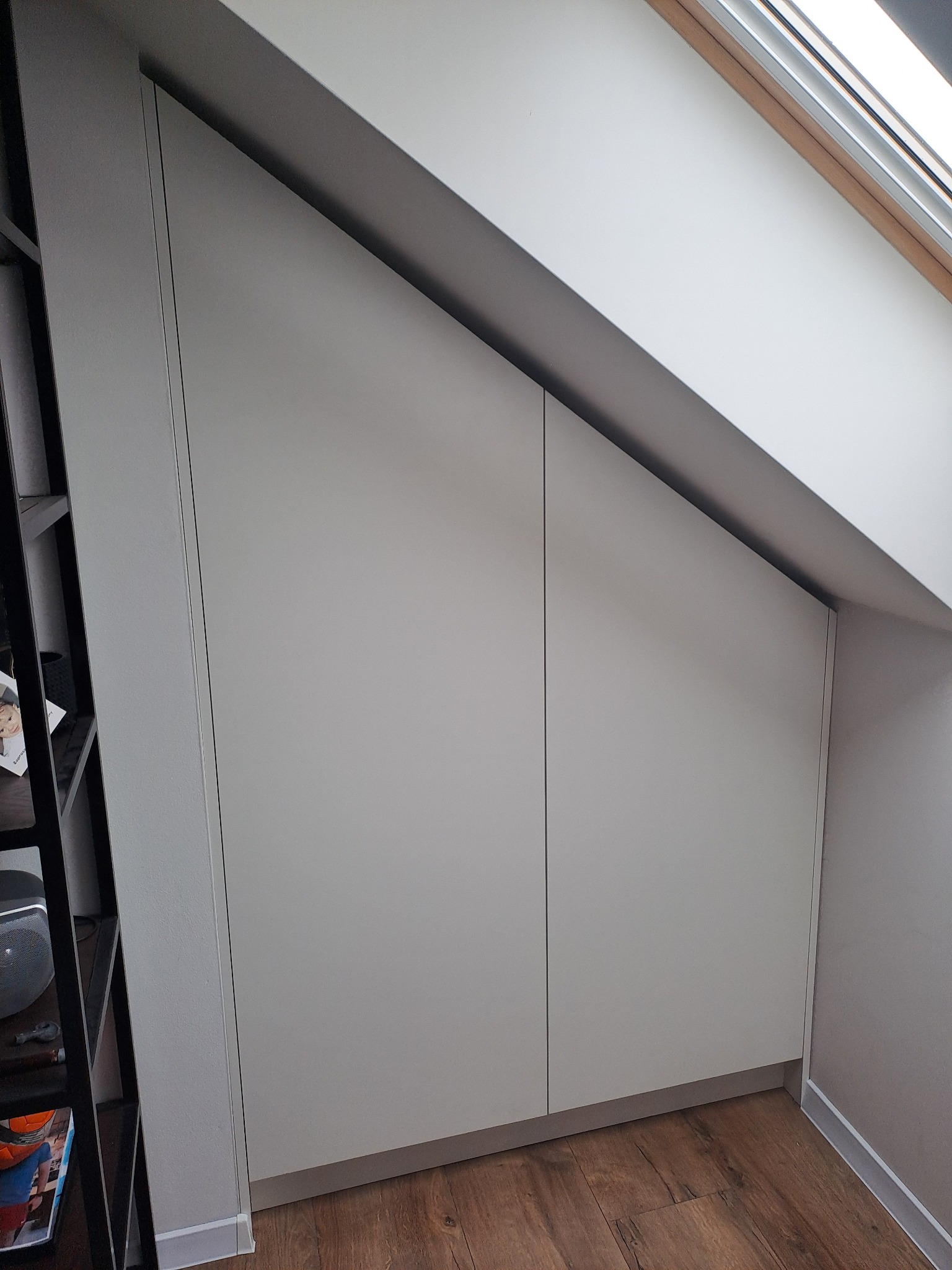 Biała szafa wnękowa na poddaszu ze skośnym sufitem, dopasowana do kąta dachu, widoczna część drewnianej podłogi i regał z książkami.