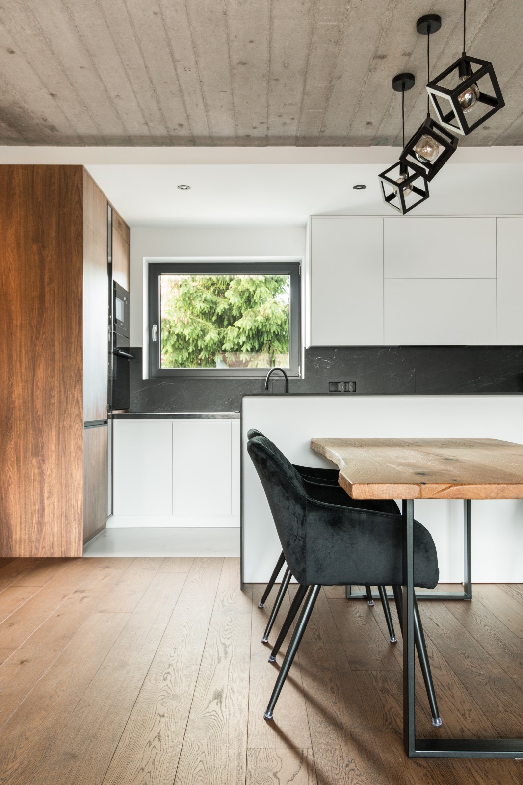 Nowoczesna kuchnia z drewnianymi frontami, białymi szafkami, blatem z czarnego kamienia i drewnianym stołem z czarnymi krzesłami na tle betonowego sufitu i parkietu.