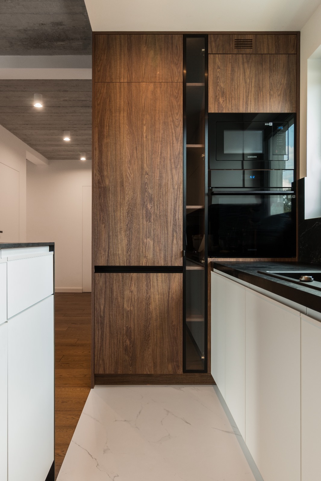 Nowoczesna kuchnia z frontami z ciemnego drewna, czarnymi akcentami i białymi szafkami, oświetlona punktowymi lampami podsufitowymi.