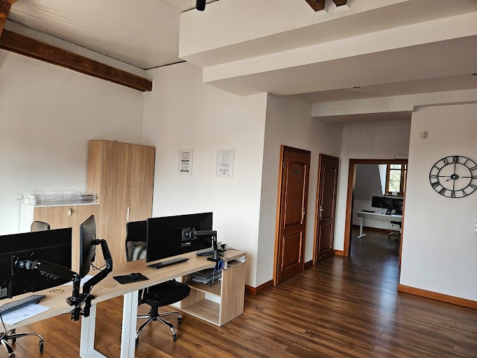 Przestronne biuro z dwoma stanowiskami pracy wyposażonymi w monitory, biurka i krzesła obrotowe, widok na dalsze pomieszczenia i duży zegar na ścianie.