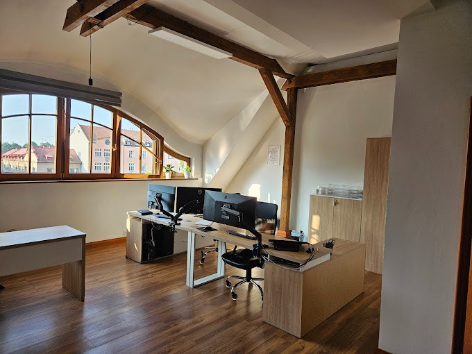 Biuro w adaptowanym poddaszu z drewnianą konstrukcją stropu, jasnymi ścianami i parkietem. Stanowiska pracy z komputerami i krzesłami biurowymi. Widok na miasto przez okna dachowe.