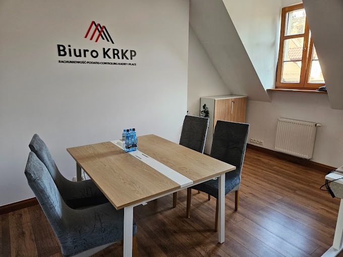 Wnętrze biura z drewnianą podłogą, stołem z czterema krzesłami i logo firmy na ścianie. Na stole stoją butelki wody.