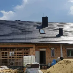 Nowo położony, ciemny dach z dachówki ceramicznej na surowym budynku z cegły, widoczny komin, okno dachowe, rynna i instalacja odgromowa, na pierwszym planie piasek i materiały budowlane.