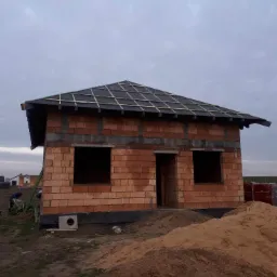 Surowy budynek jednorodzinny z częściowo wykonanym dachem, widoczne łaty pod pokrycie i cegły ceramiczne, na pierwszym planie hałda piasku.