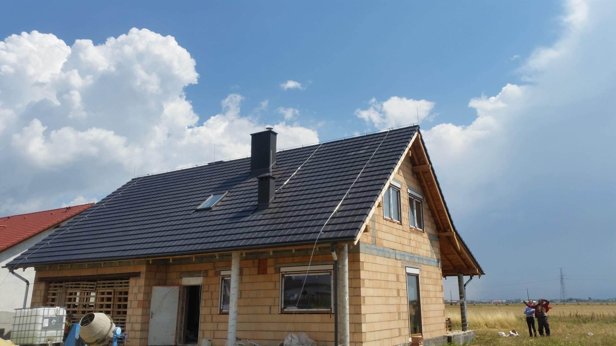 Dom w stanie surowym zamkniętym z nowym, ciemnym dachem i kominem na tle błękitnego nieba z obłokami.
