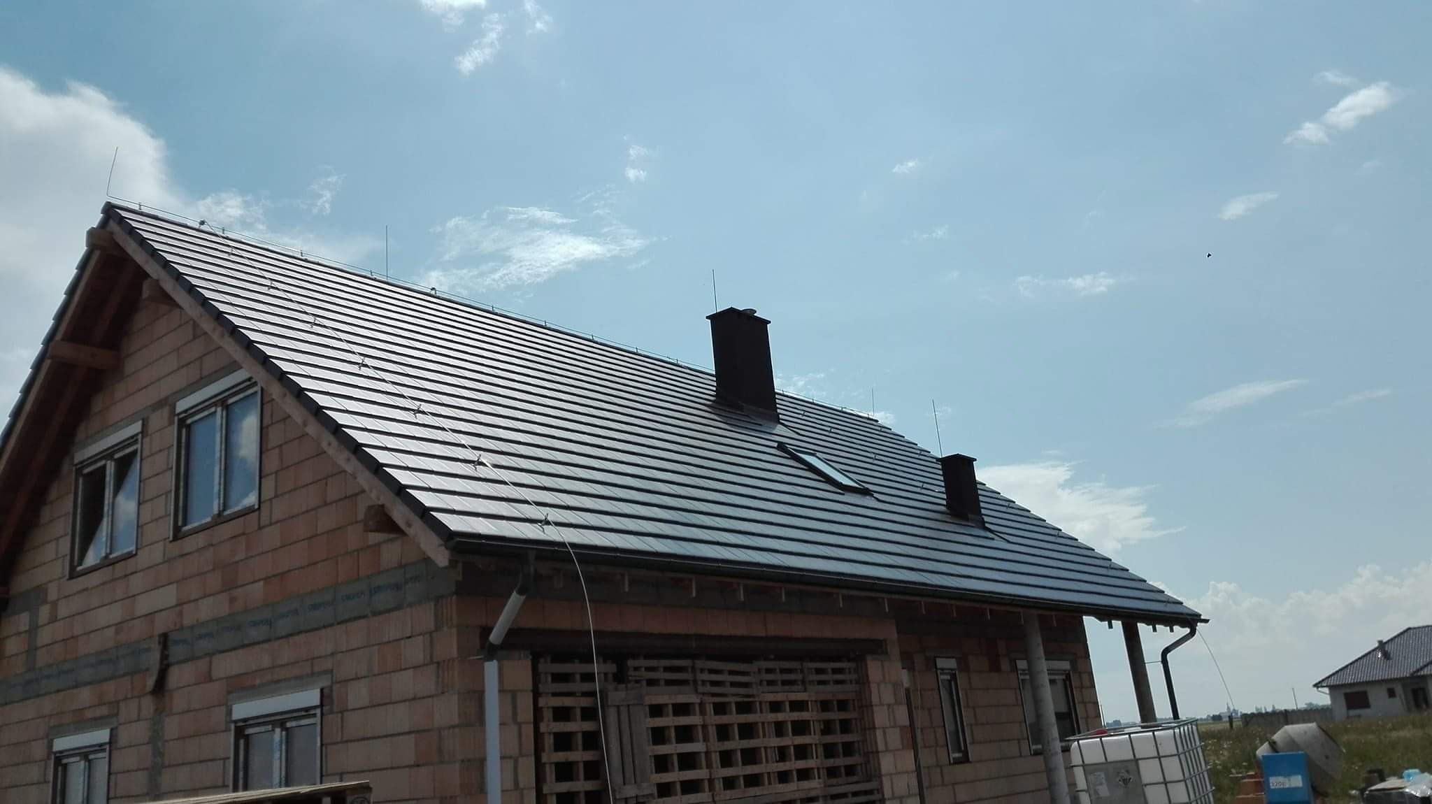 Dom jednorodzinny w trakcie budowy z nowym, ciemnym dachem z dachówki ceramicznej, widoczne kominy i okno dachowe, orynnowanie oraz niezakończona elewacja z cegły kratówki
