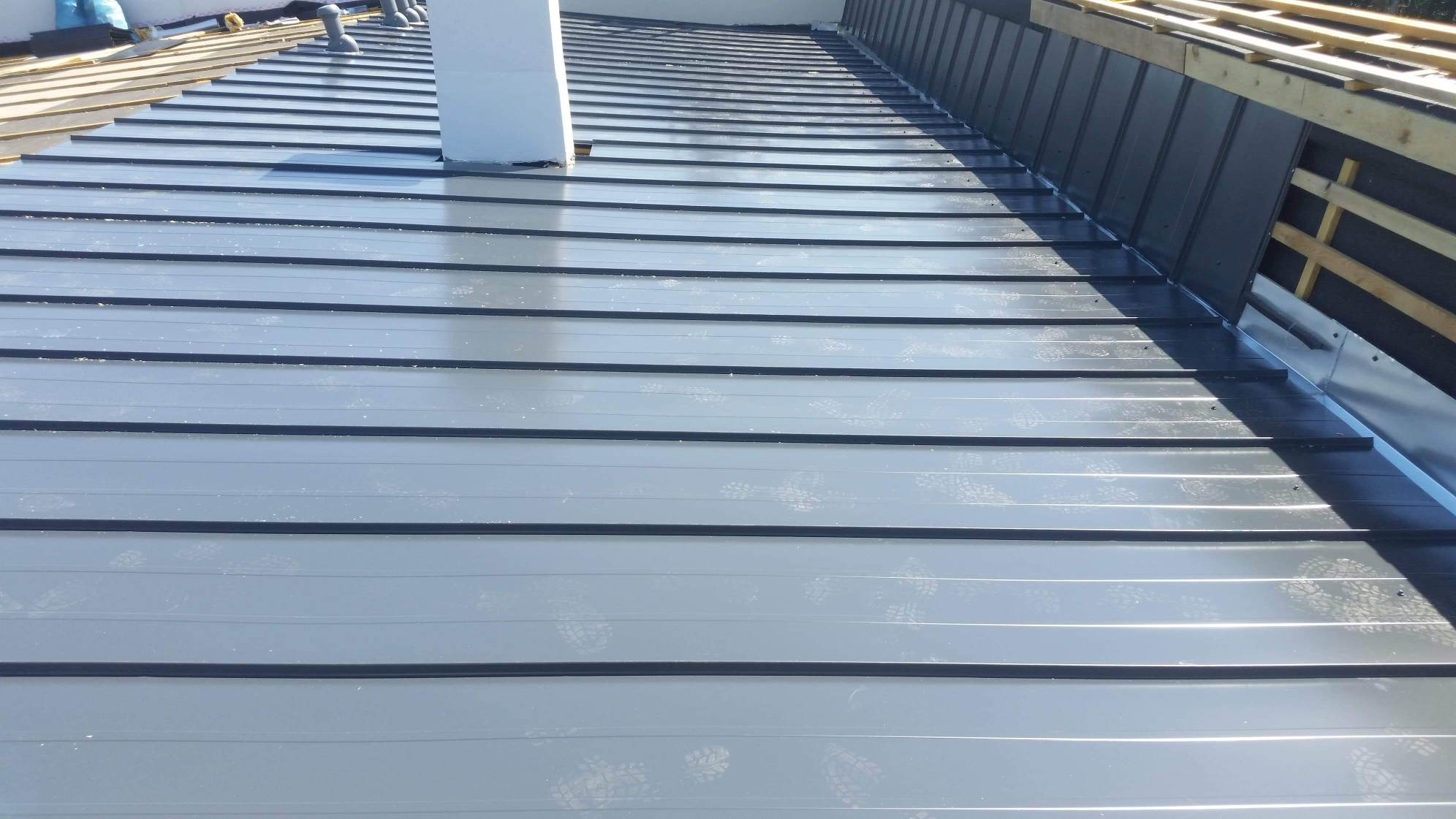 Szare blaszane pokrycie dachowe z widocznymi odciskami butów, kominem i fragmentem drewnianej konstrukcji dachu w trakcie montażu.