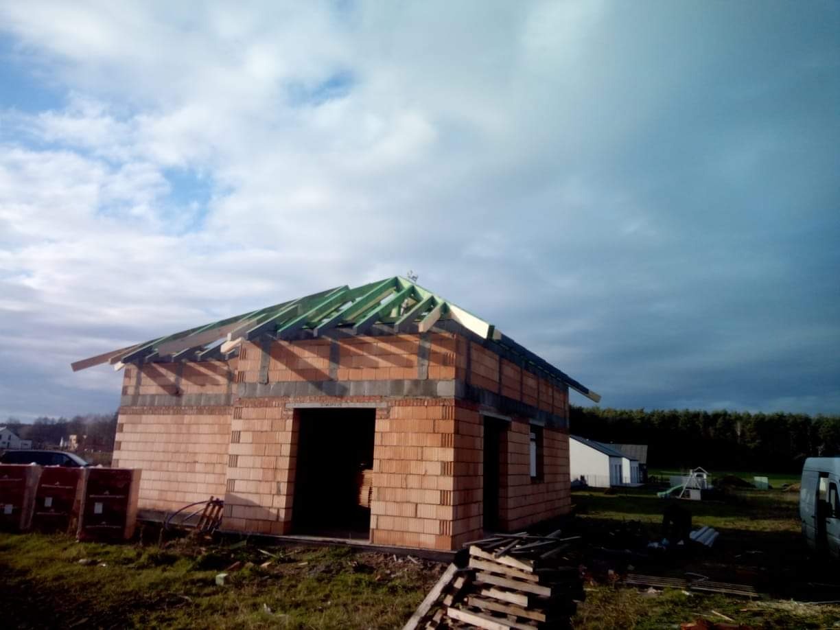 Budowa domu jednorodzinnego w stanie surowym otwartym: ściany z czerwonej cegły, zielona konstrukcja drewniana dachu w trakcie montażu, widoczne palety z cegłami i elementy konstrukcyjne...