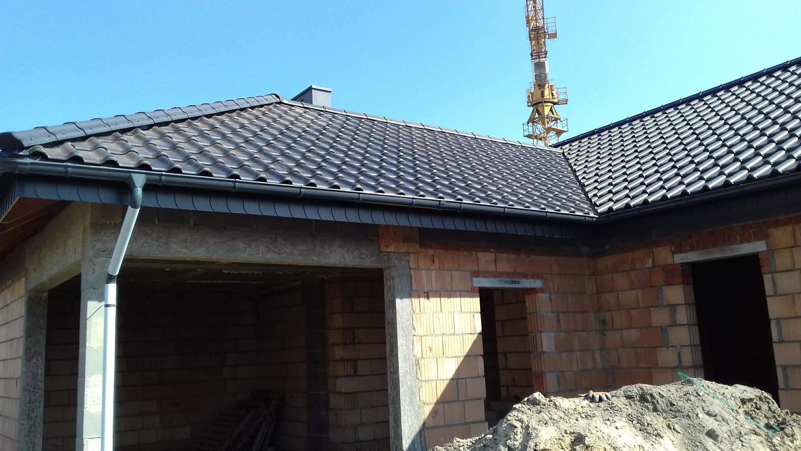 Nowy dach z ciemnej dachówki ceramicznej i orynnowaniem zamontowany na surowym budynku w trakcie budowy, widoczny żuraw budowlany w tle.
