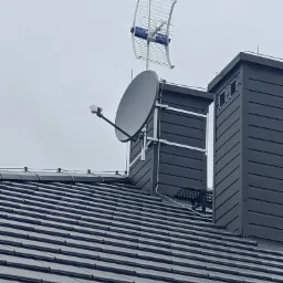 Antena satelitarna i antena naziemna zamontowane na kominie domu z ciemną dach&oacute;wką, widoczne elementy mocujące.