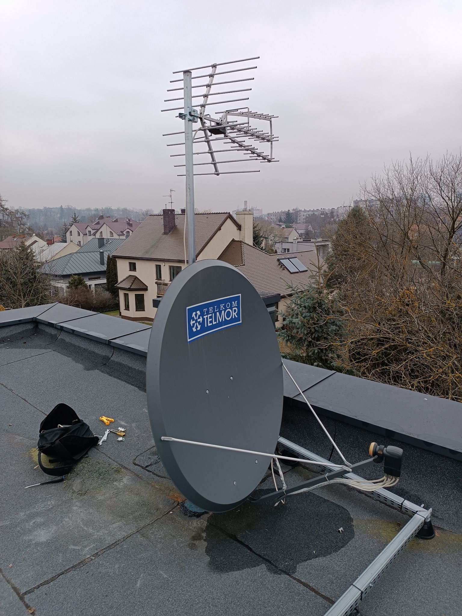 Antena satelitarna Telkom Telmor i antena kierunkowa Yagi-Uda zamontowane na płaskim dachu budynku, widoczne narzędzia i torba monterska, w tle zabudowa miejska w pochmurny dzień.