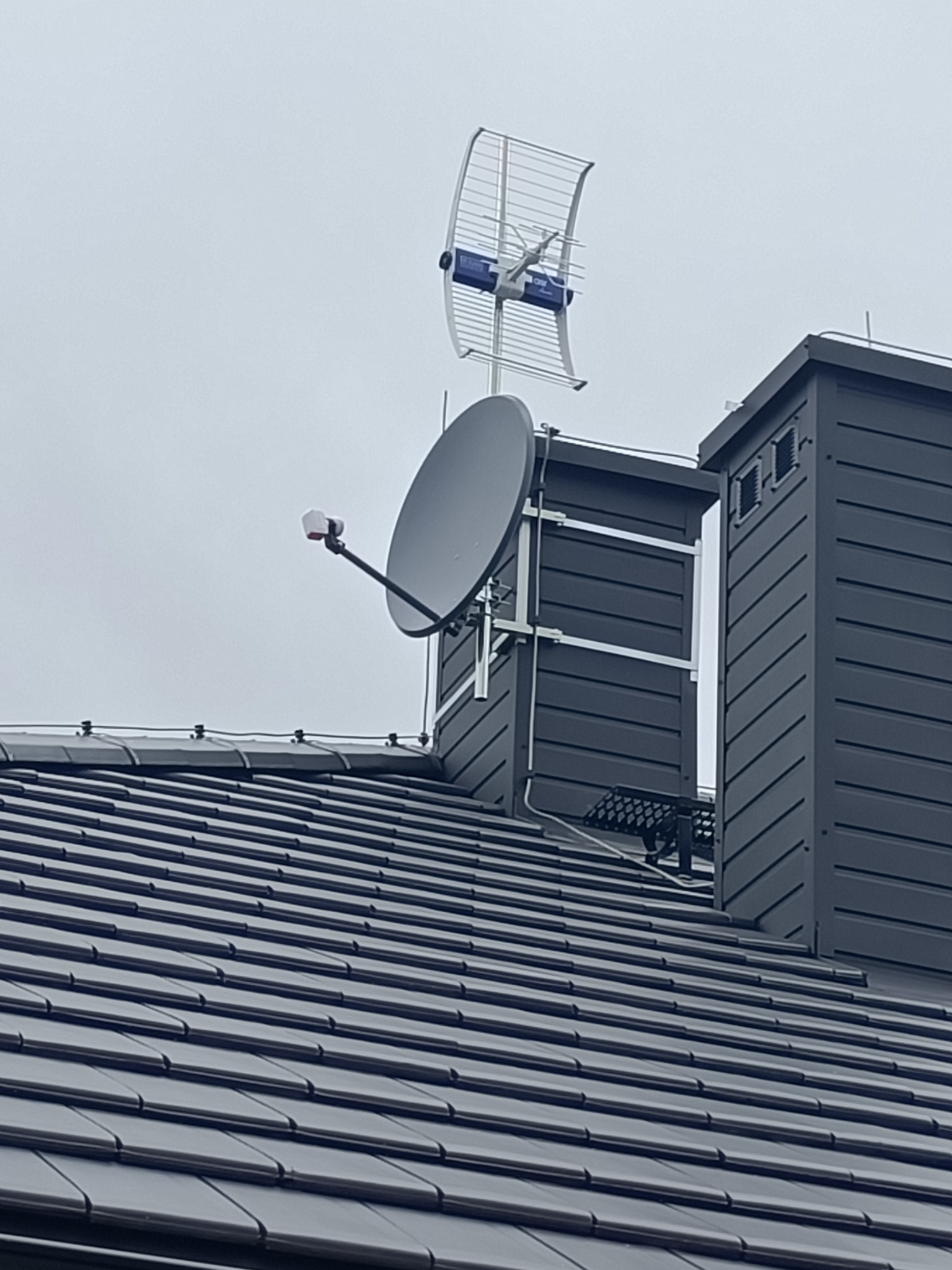 Antena satelitarna i antena naziemna zamontowane na kominie domu z ciemną dachówką, widoczne elementy mocujące.
