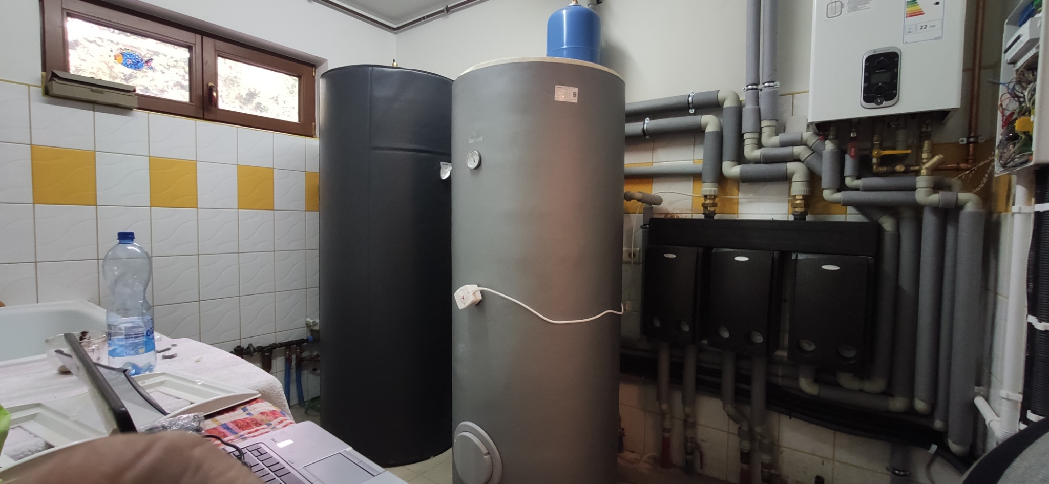 Dwa zbiorniki buforowe, czarny i szary, stojące obok siebie w kotłowni z białymi i żółtymi kafelkami, widoczne rury i elementy instalacji grzewczej, oraz fragment laptopa w dolnym rogu.