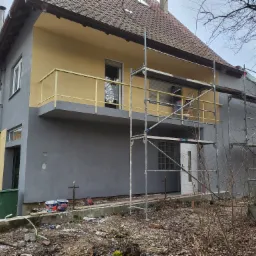 Dwukondygnacyjny dom z częściowo pomalowaną elewacją na szaro i żółto, widoczne rusztowania, puszki z farbą przy fundamencie i pracownik w trakcie malowania na balkonie.