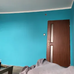 Pomieszczenie po malowaniu ścian na turkusowo, widoczna drewniana ościeżnica drzwiowa z szybą, zabezpieczone meble i akcesoria malarskie na stole.