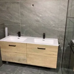 Nowoczesna łazienka z podwójną umywalką na szafce z jasnego drewna, czarne baterie, szare płytki na ścianach i podłodze, widoczna kabina prysznicowa.