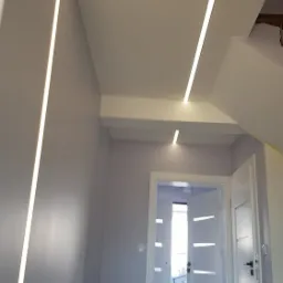 Nowoczesne oświetlenie LED wpuszczone w sufit, tworzące geometryczny wzór w korytarzu z otwartymi białymi drzwiami.