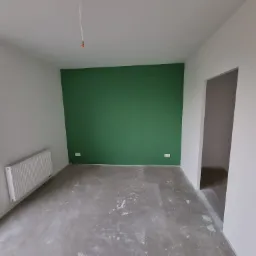 Pusty pokój po malowaniu: jedna ściana pomalowana na intensywny zielony kolor, pozostałe ściany białe, szara betonowa podłoga, biały grzejnik pod oknem, wystające kable ze ściany