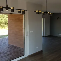 Nowoczesne oświetlenie w przestronnym wnętrzu z drewnianą podłogą i ścianą z dekoracyjnej cegły, widok na taras z huśtawką.