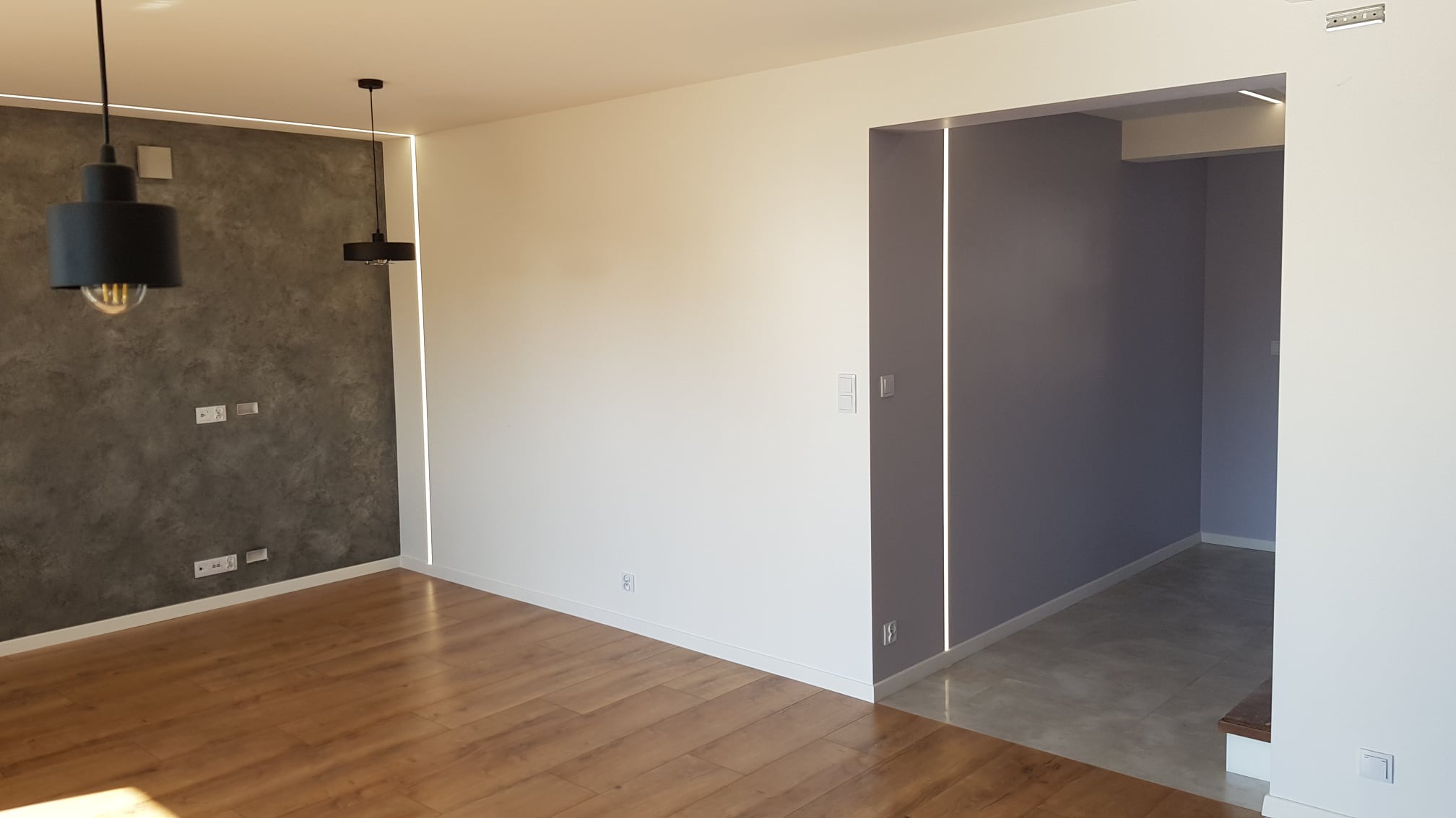 Nowoczesne wnętrze: pokój z drewnianą podłogą, ściana pokryta szarą dekoracyjną farbą, dwie wiszące lampy z odsłoniętymi żarówkami, białe ściany z listwami LED wzdłuż sufitu i przejścia...