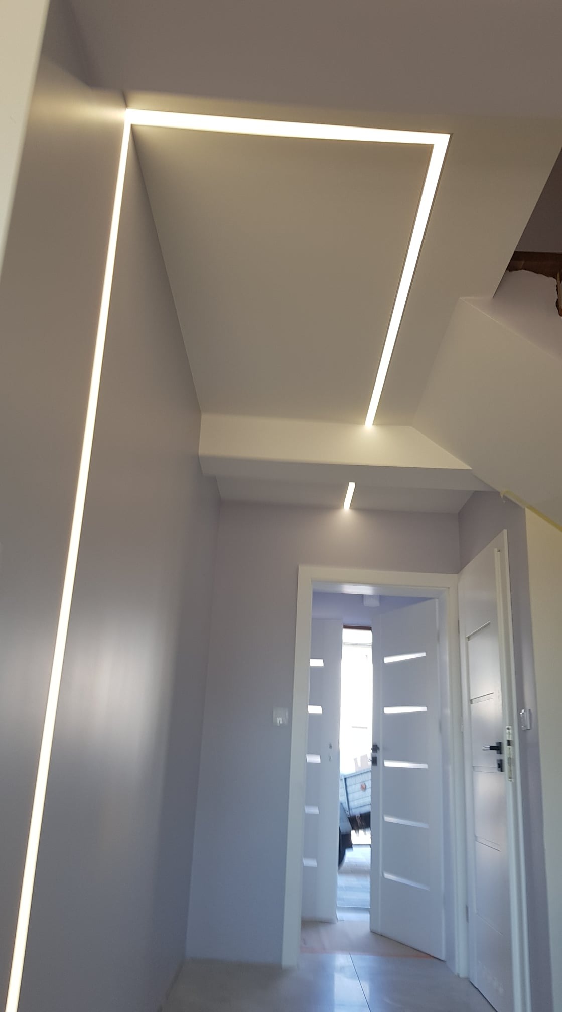 Nowoczesne oświetlenie LED wpuszczone w sufit, tworzące geometryczny wzór w korytarzu z otwartymi białymi drzwiami.