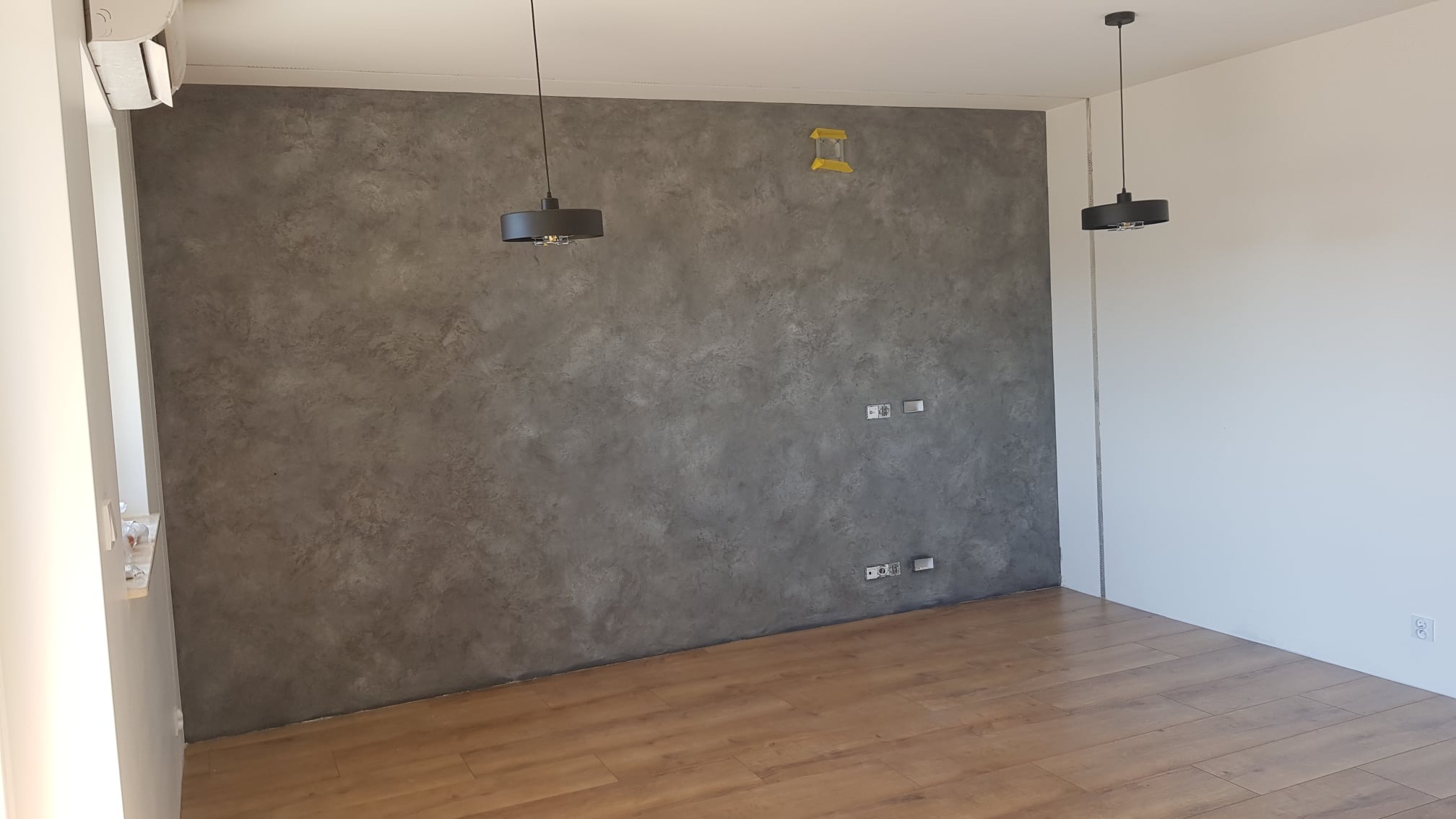 Wnętrze pokoju z dekoracyjną ścianą w odcieniach szarości, drewnianą podłogą, dwoma nowoczesnymi lampami wiszącymi i klimatyzatorem przy suficie.