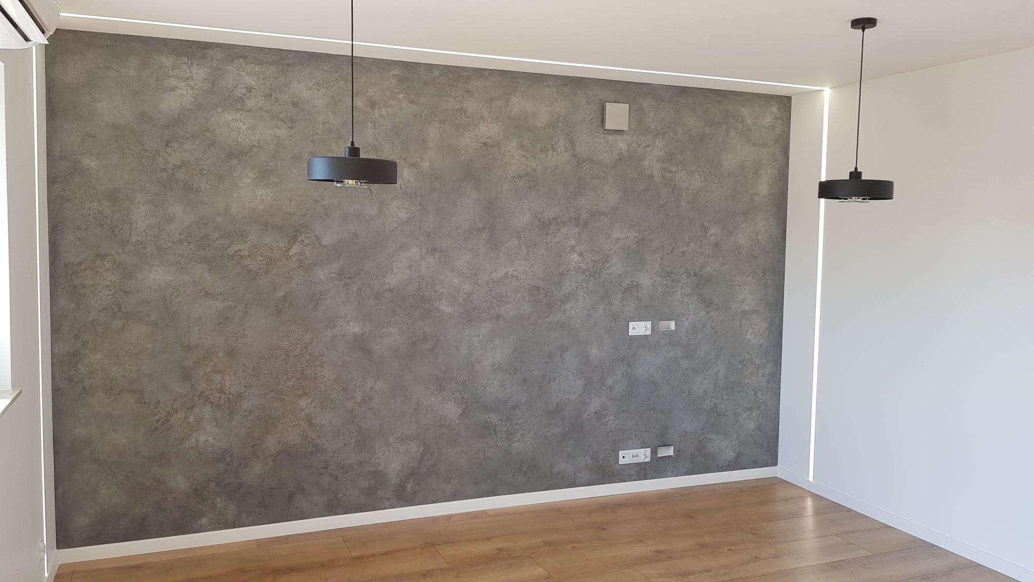 Szara ściana z dekoracyjną fakturą, oświetlona liniowym światłem LED w suficie i dwoma wiszącymi lampami z czarnymi abażurami, parkiet na podłodze.