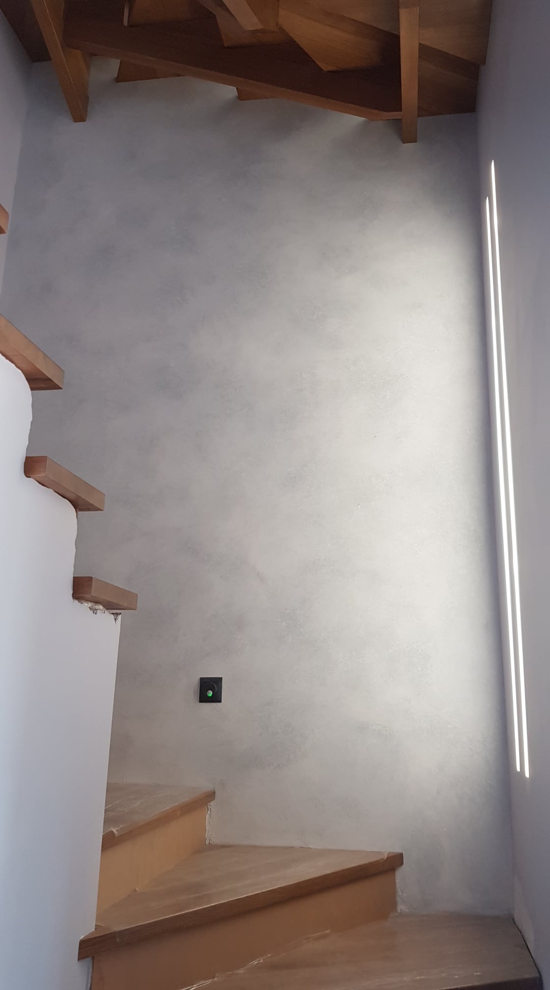 Schody drewniane z dekoracyjną szarą ścianą o fakturze betonu i podłużnym oświetleniem LED wzdłuż krawędzi ściany, widoczne gniazdko elektryczne.