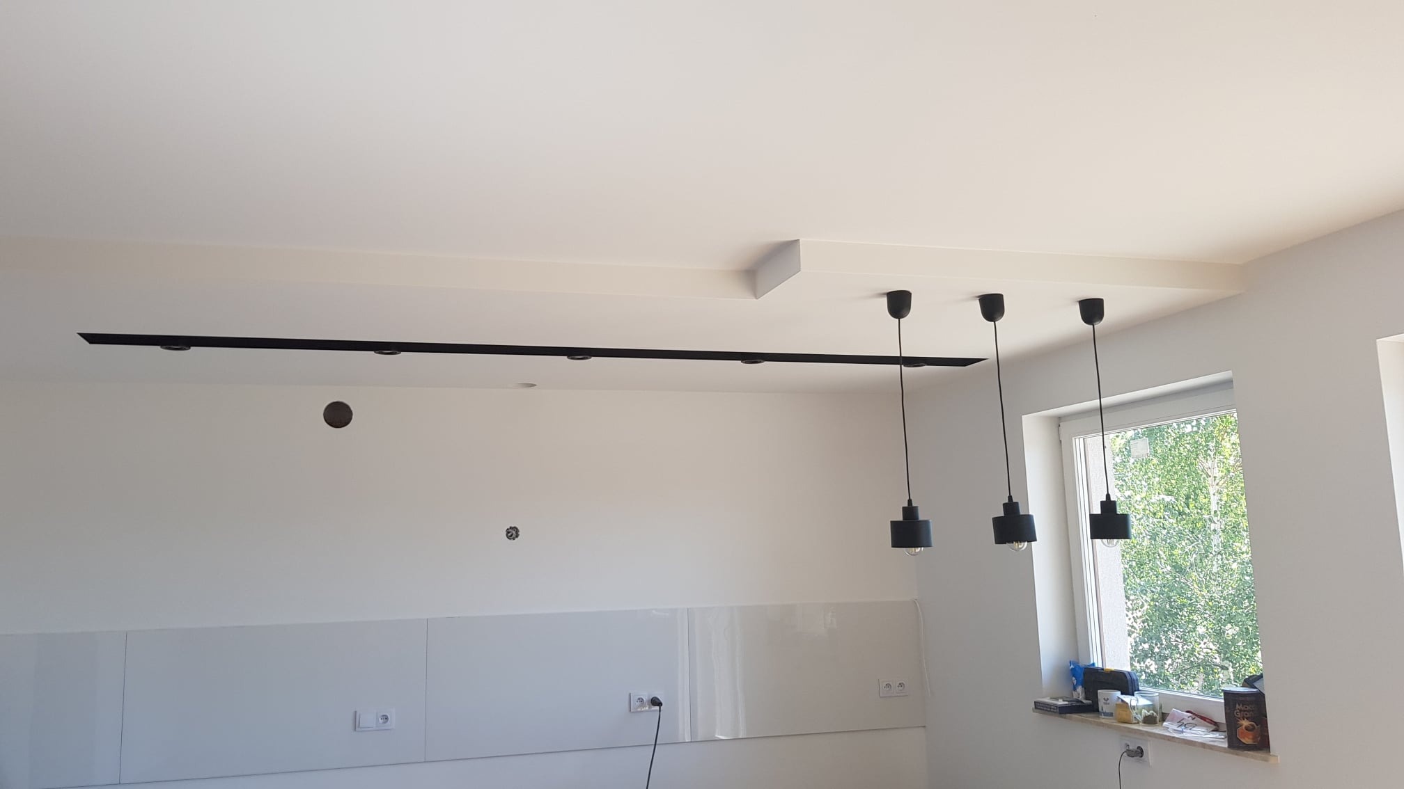 Białe wnętrze w trakcie wykańczania: zamontowana czarna szyna oświetleniowa z reflektorami na suficie, trzy czarne lampy wiszące, widoczne puszki elektryczne na ścianie, okno z widokiem na zieleń.