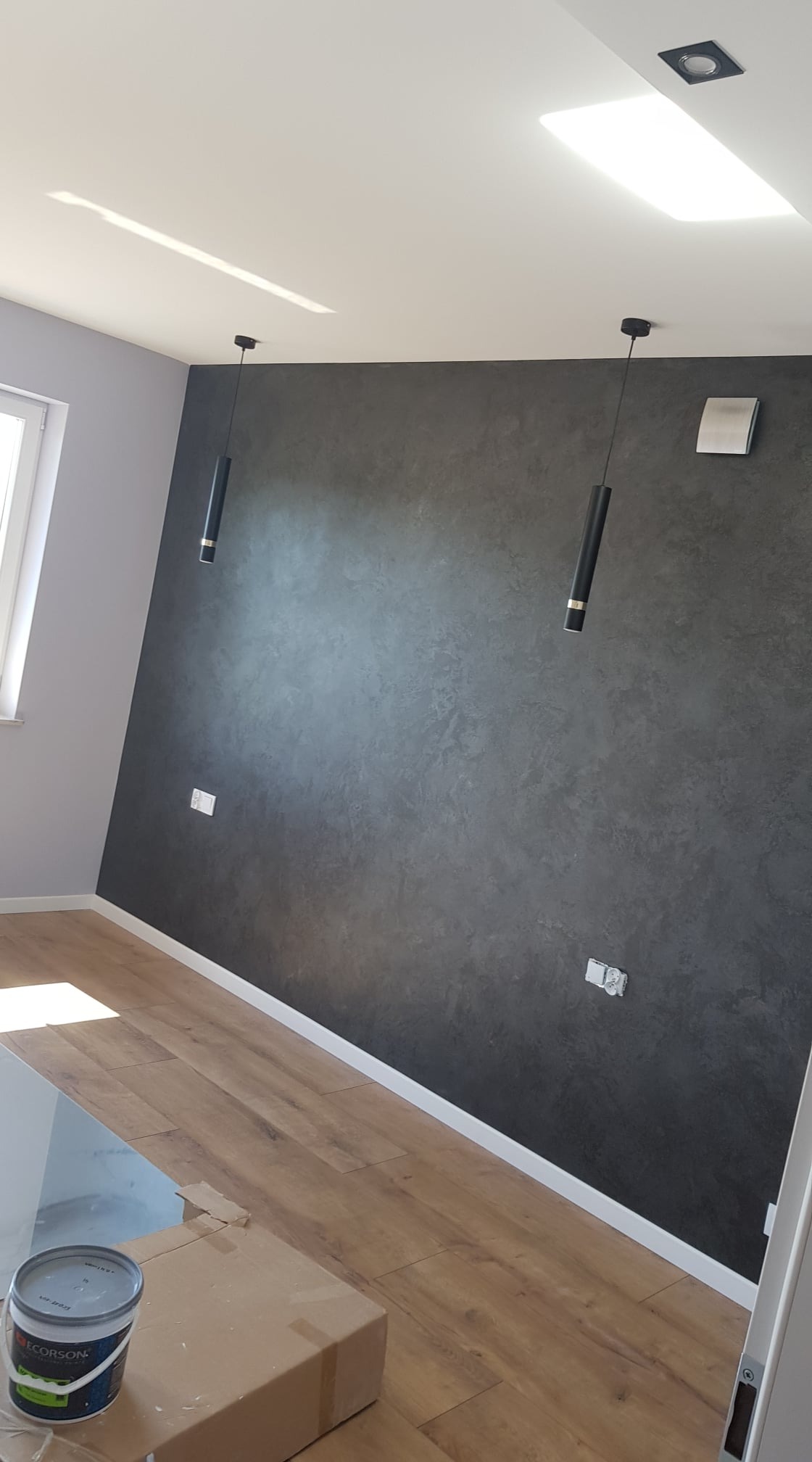 Pokój po malowaniu z efektem dekoracyjnym na ścianie, widoczne dwie wiszące lampy tuby, drewnopodobna podłoga i puszka farby na kartonie.