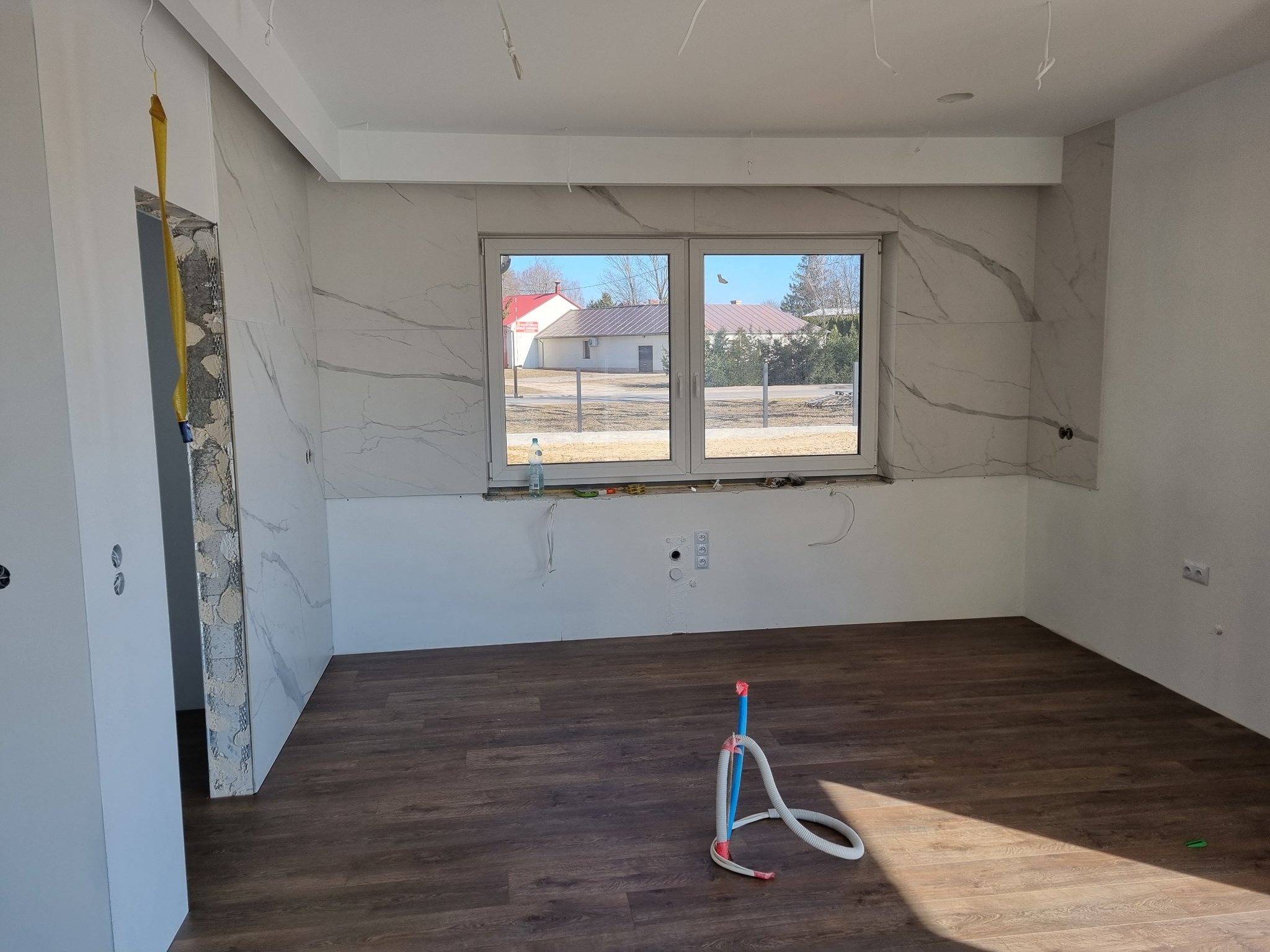 Wnętrze pokoju w trakcie prac wykończeniowych; widoczne płytki imitujące marmur na ścianach, świeżo położona podłoga z paneli, wystające kable i rury, oraz fragment okna z widokiem na budynki.