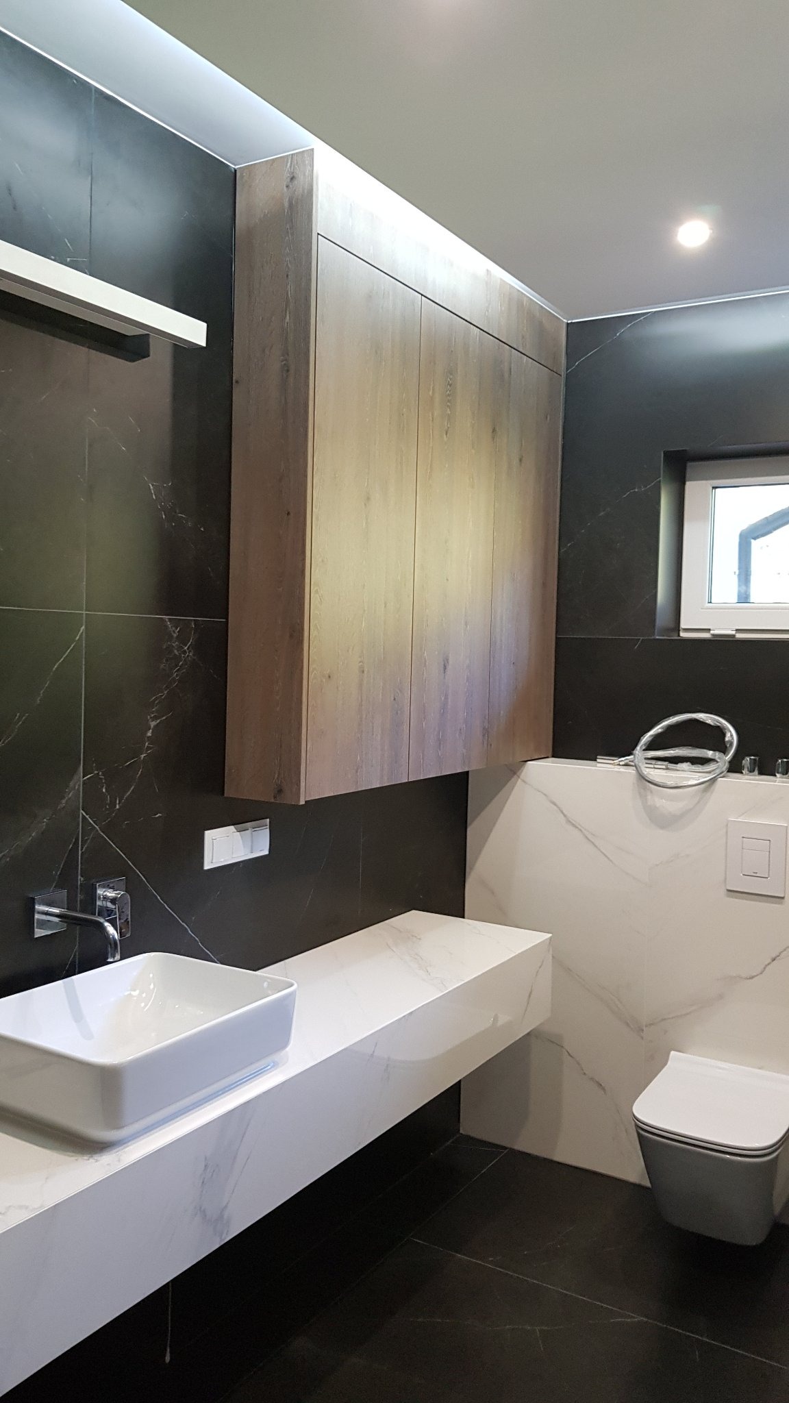 Nowoczesna łazienka z czarnymi marmurowymi płytkami, białym blatem umywalkowym z kwadratową umywalką, drewnianą szafką wiszącą i szarą podwieszaną toaletą.