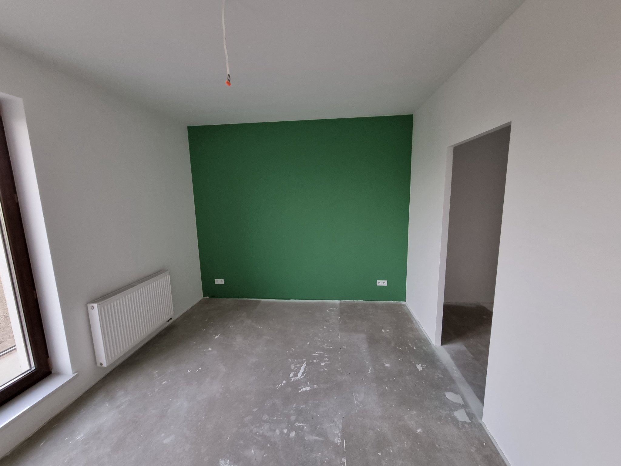 Pusty pokój po malowaniu: jedna ściana pomalowana na intensywny zielony kolor, pozostałe ściany białe, szara betonowa podłoga, biały grzejnik pod oknem, wystające kable ze ściany