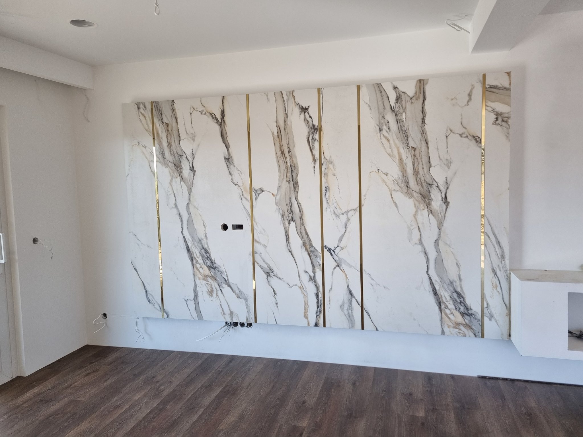 Ściana z paneli imitujących marmur, oddzielonych złotymi listwami, widoczne otwory na gniazdka elektryczne i wystające kable, podłoga z paneli drewnopodobnych.