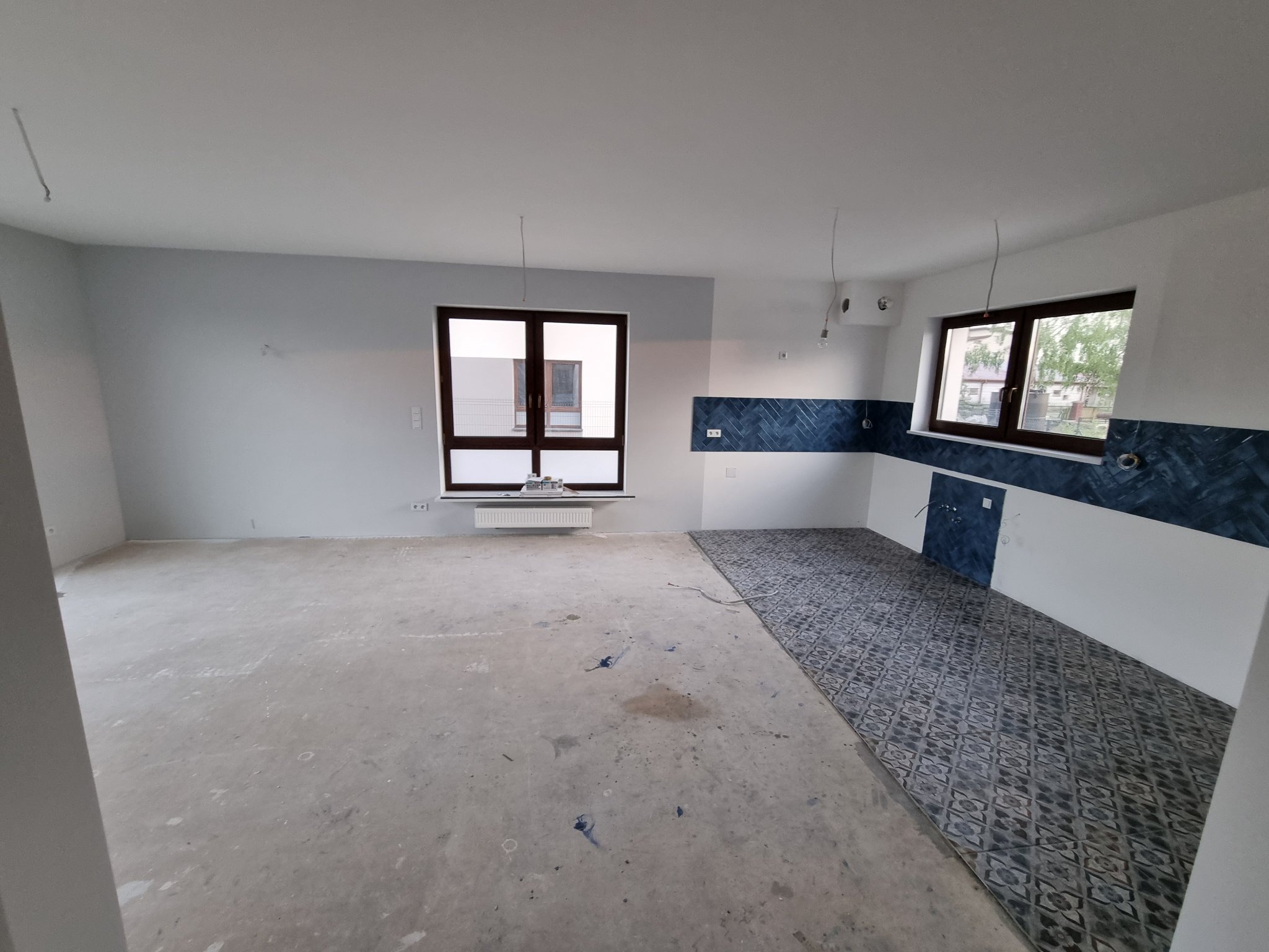 Wnętrze w trakcie remontu: częściowo pomalowane ściany na szaro, fragment ułożonej podłogi z wzorzystych płytek w odcieniach szarości i błękitu, dwa okna z widokiem na zewnątrz.
