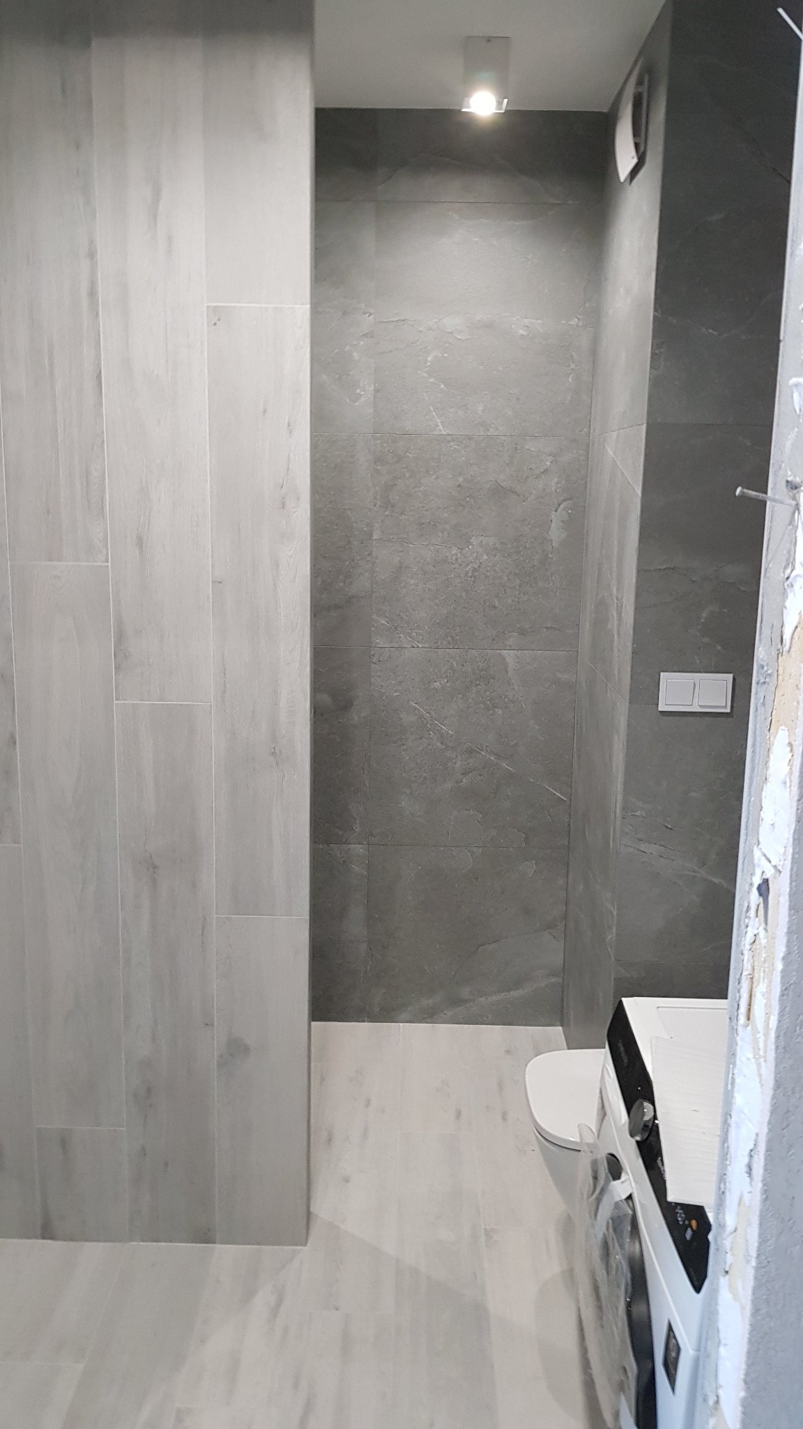 Nowoczesna łazienka z szarymi płytkami imitującymi kamień na ścianach i podłodze, biała armatura i pralka w folii ochronnej, minimalistyczne oświetlenie punktowe.