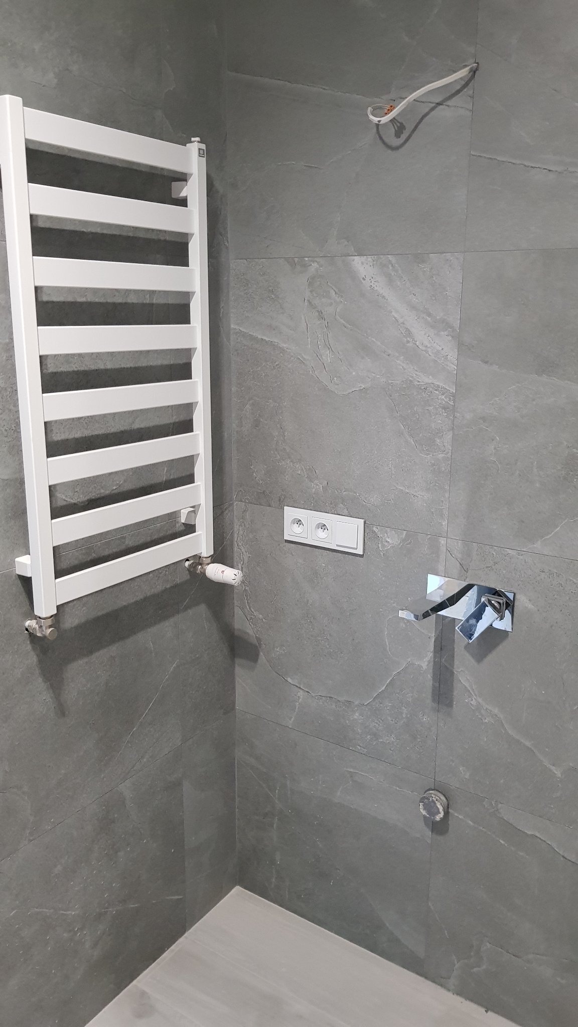Szara łazienka w trakcie wykańczania: biały grzejnik drabinkowy, nowoczesna bateria umywalkowa, podwójne gniazdko elektryczne, wystający przewód elektryczny, szare płytki imitujące kamień.