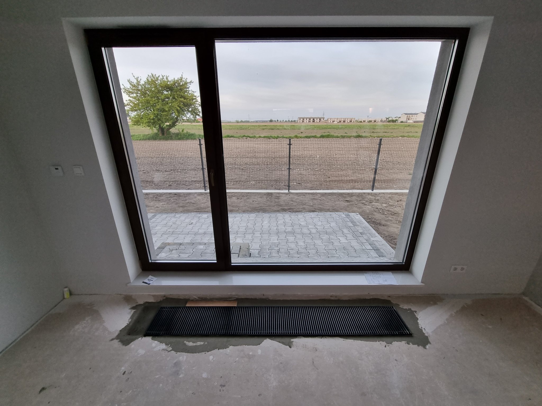 Wnętrze w trakcie remontu w Białej Podlaskiej: widok z okna na zewnątrz, gdzie widać świeżo ułożoną kostkę brukową i pole. Na podłodze wylewka i zamontowany grzejnik kanałowy.